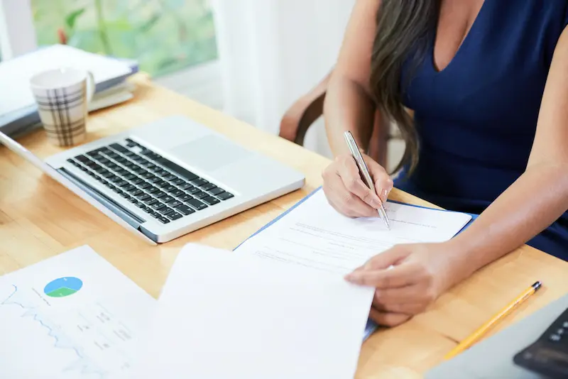 Woman signing documents at her desk next to a laptop.